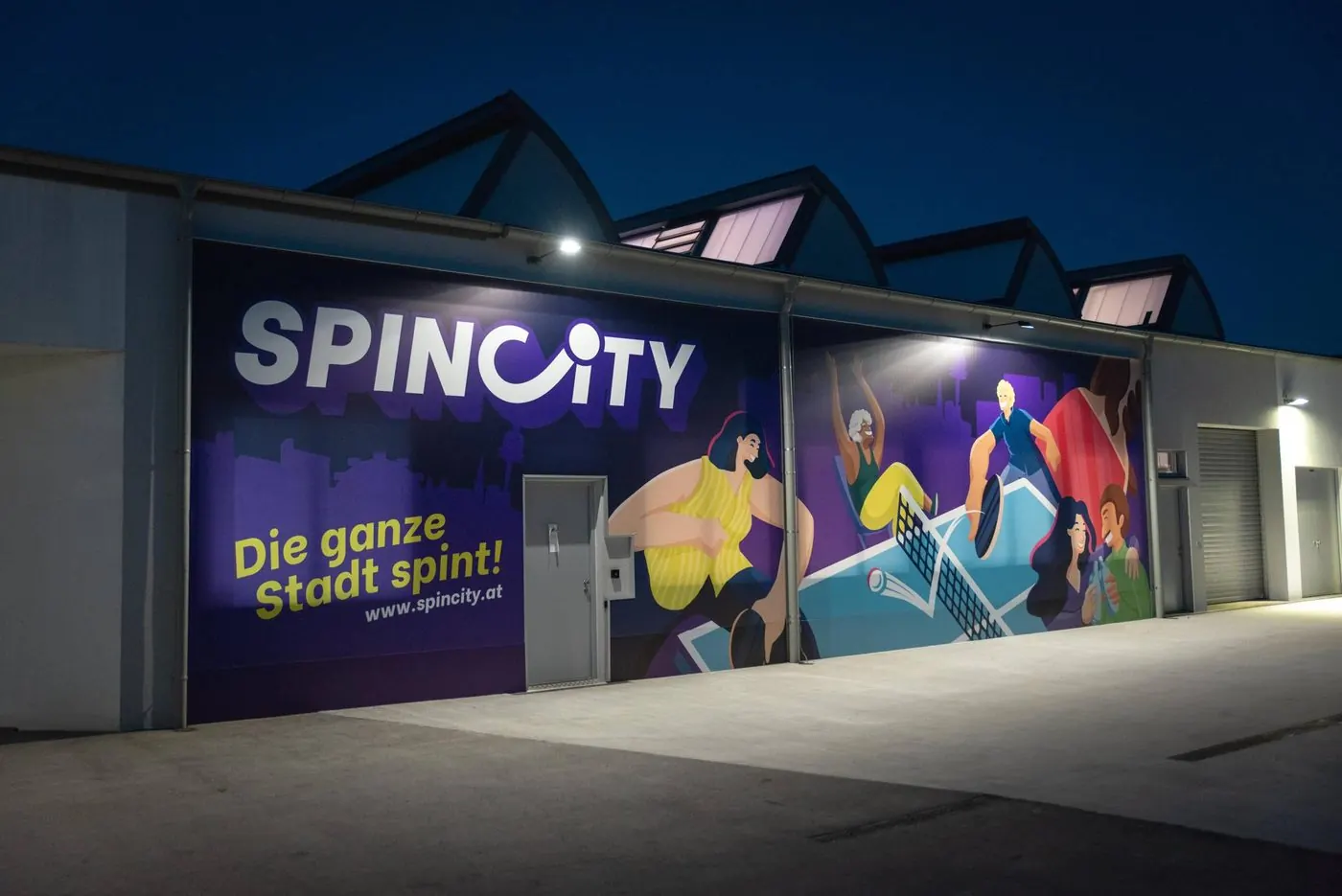 SpinCity from outside in the evening: grey hall with a large purple mural “SpinCity – Die ganze Stadt spint!” and the entrance door