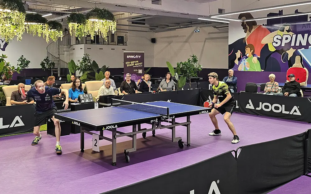 Meisterschaftsspiel in der SpinCity: zwei Spieler am Tisch, Zuschauer in der Lounge