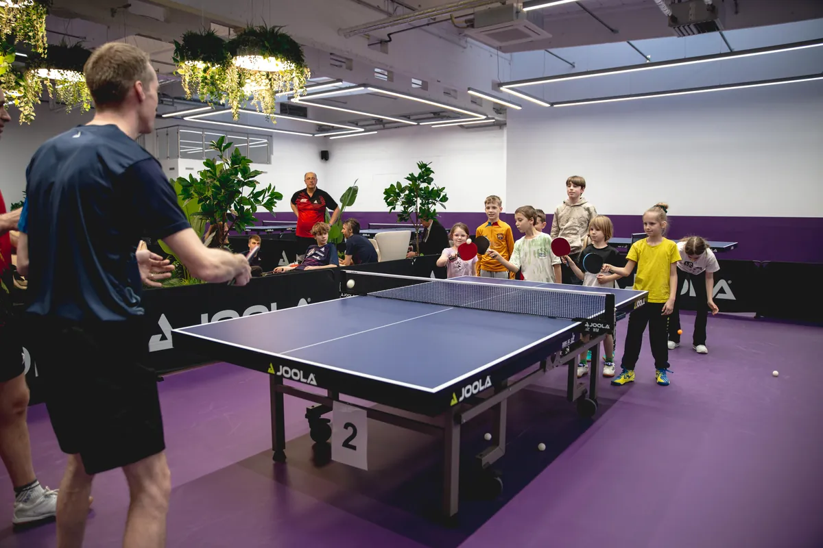 Kinder warten mit Schlägern an einem Tischtennistisch, vorne spielt ein Betreuer den Ball zu